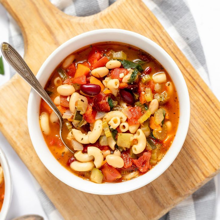 a white bowl filled with pasta soup on top of a wooden cutting board next to a spoon