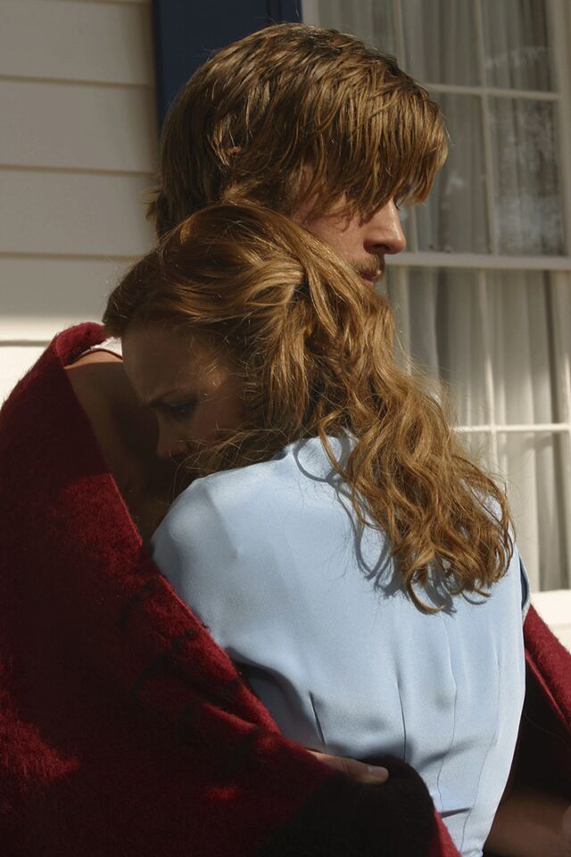 a woman wrapped in a red blanket standing next to a man with long curly hair