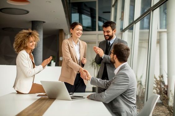 three business people shaking hands in front of a laptop on a table with two other people