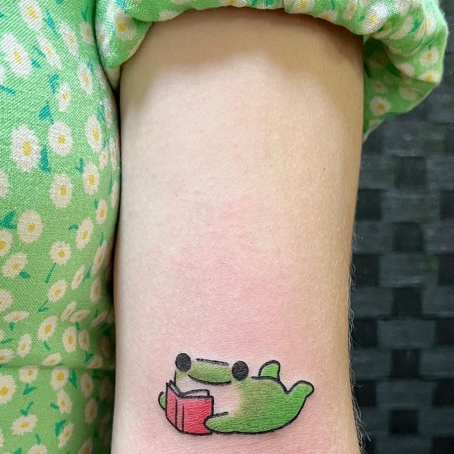 a small frog reading a book tattoo on the left side of the arm, with daisies in the background