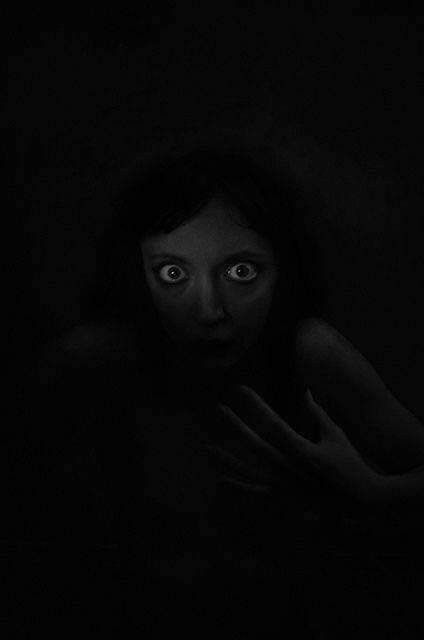 a woman is hiding in the dark with her hands on her chest and looking at the camera