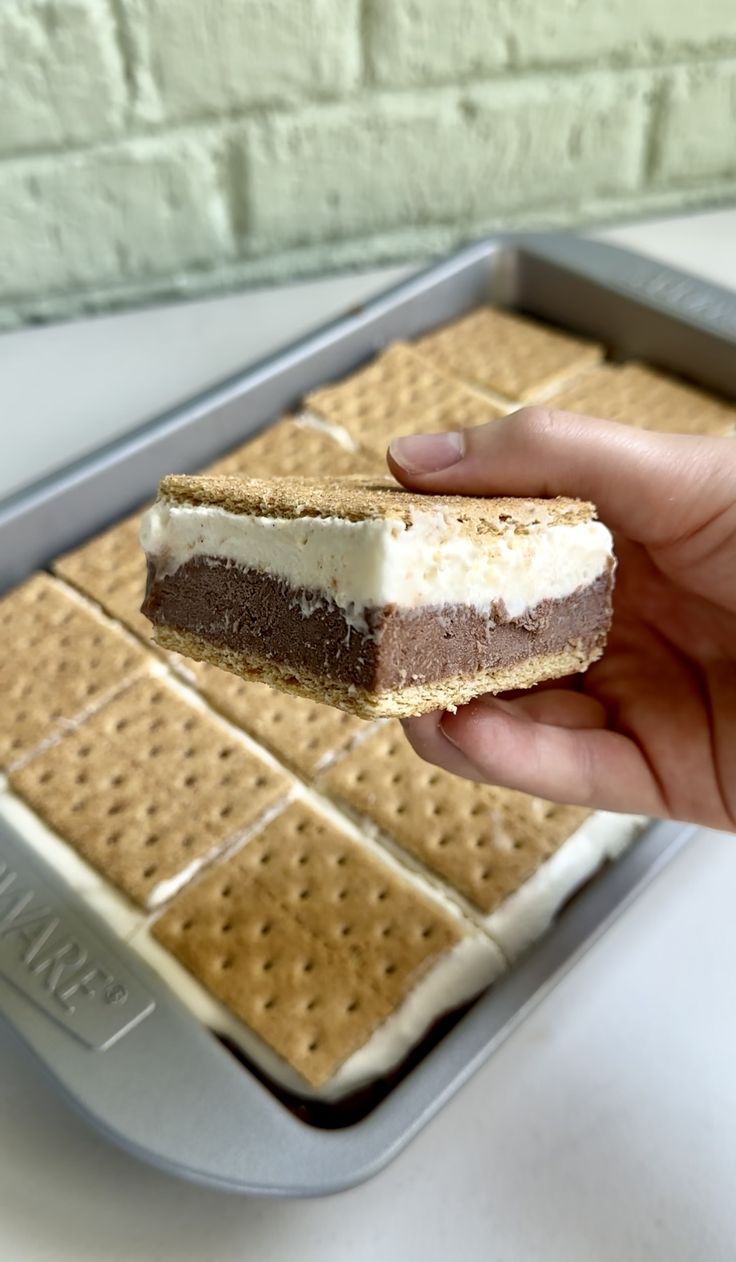 a hand holding a piece of cake on top of a cookie sheet covered in graham crackers