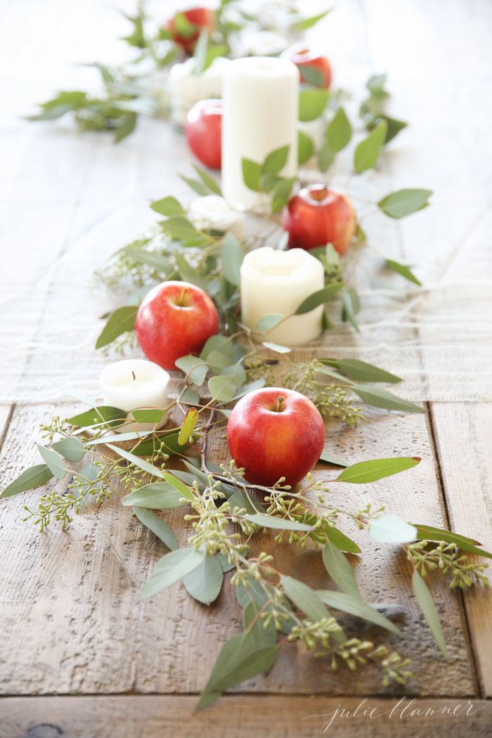 an arrangement of apples and greenery on a wooden table with candles in the background