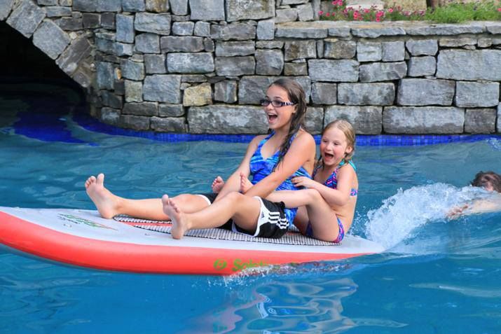 two girls are riding on a boogie board in the pool while another girl swims behind them