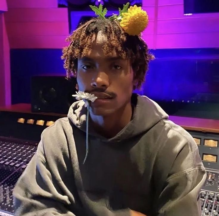 a young man sitting in front of a mixing desk with flowers on his head and tongue sticking out