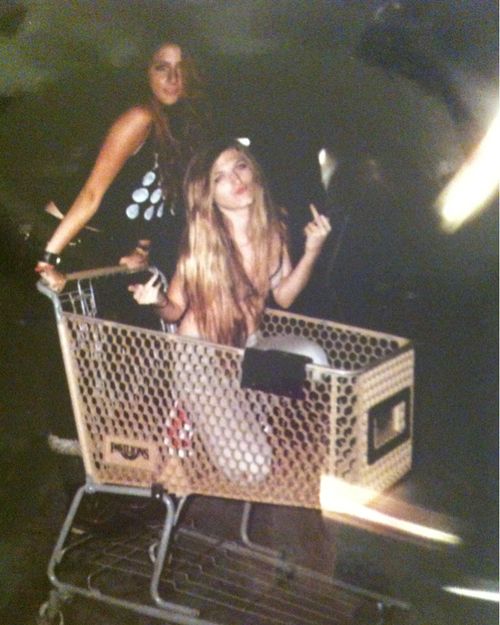 two women standing in a shopping cart with their arms up