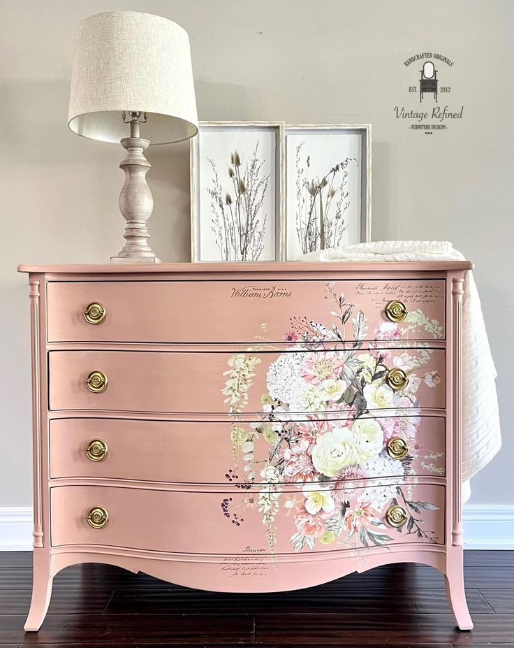 a pink dresser with flowers painted on it and two framed pictures above the drawers, along with a lamp