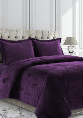 a bed with purple comforter and pillows in a white room next to a lamp