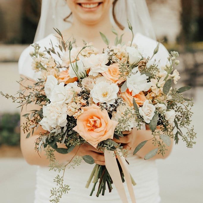 a bride holding a bouquet of white and peach flowers with greenery in her hands