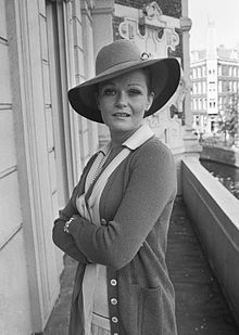 a woman in a hat and cardigan standing on a balcony with her arms crossed