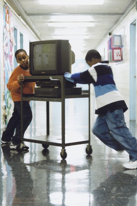 two young boys playing with an old tv on a cart in a hallway at school