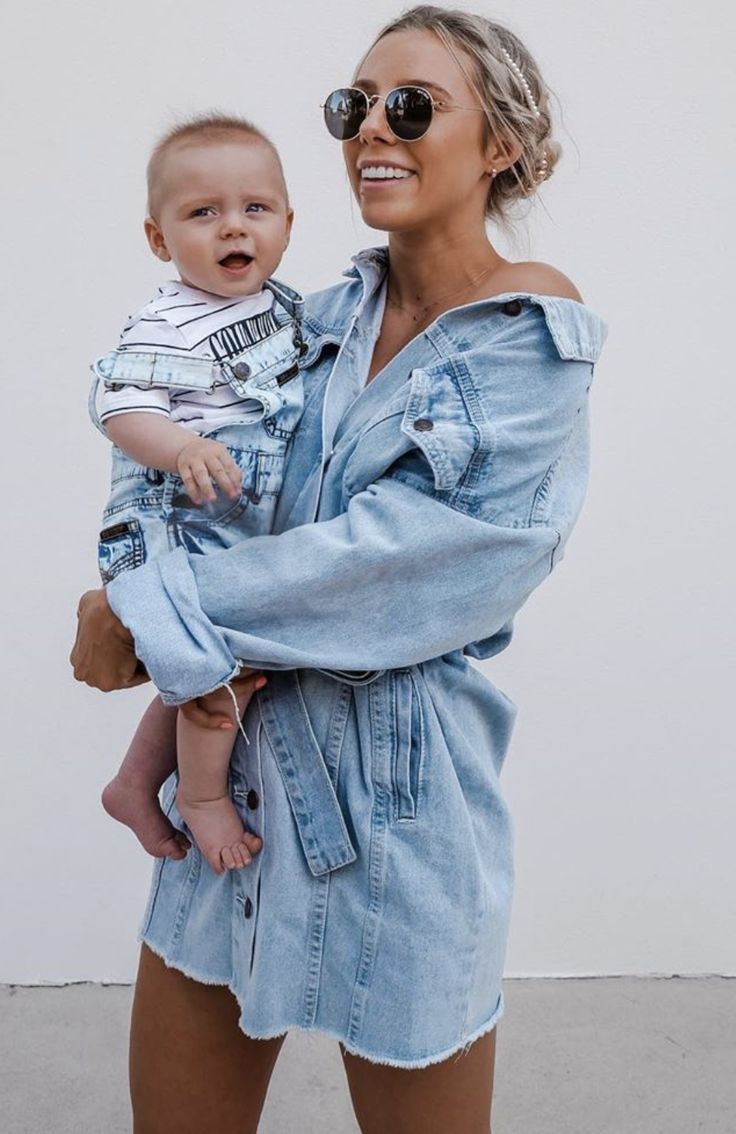 a woman holding a baby in her arms and smiling at the camera while standing next to a white wall