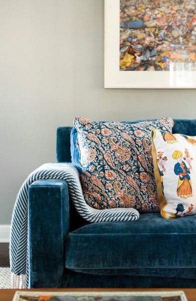 a blue velvet couch with colorful pillows on it's arms and back, in front of a framed art piece