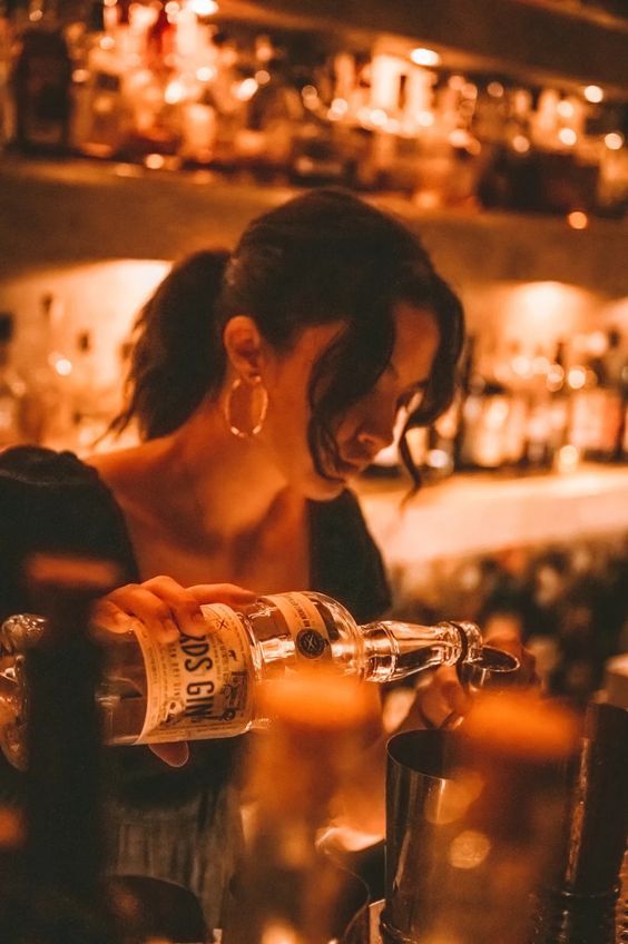 a woman pouring drinks at a bar with lots of liquor bottles on the shelves behind her