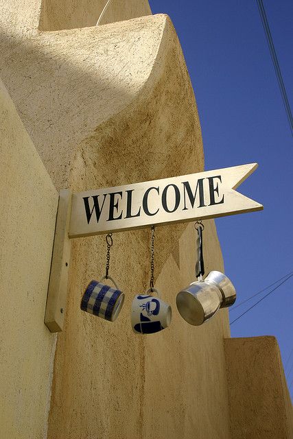 a welcome sign hanging from the side of a building with coffee cups dangling from it's hooks