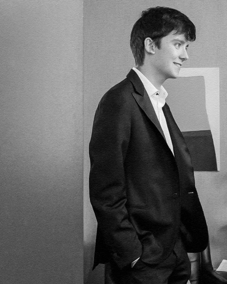 a young man in a suit and tie standing next to a desk with a computer on it