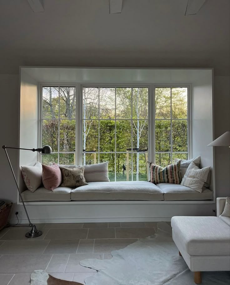 a white window seat with pillows on it in front of a large window and floor lamp