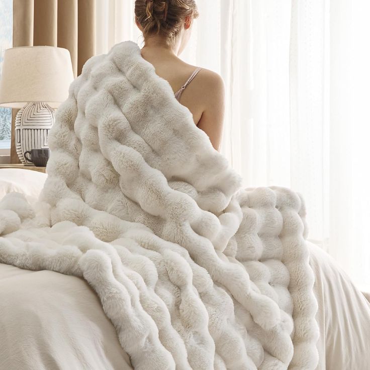 a woman sitting on a bed under a fluffy white blanket with her back turned to the camera