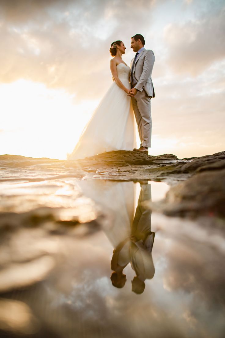 a bride and groom are standing on the rocks in front of the ocean at sunset