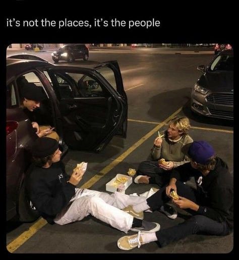 four people sitting on the ground eating hotdogs next to a car and another person standing