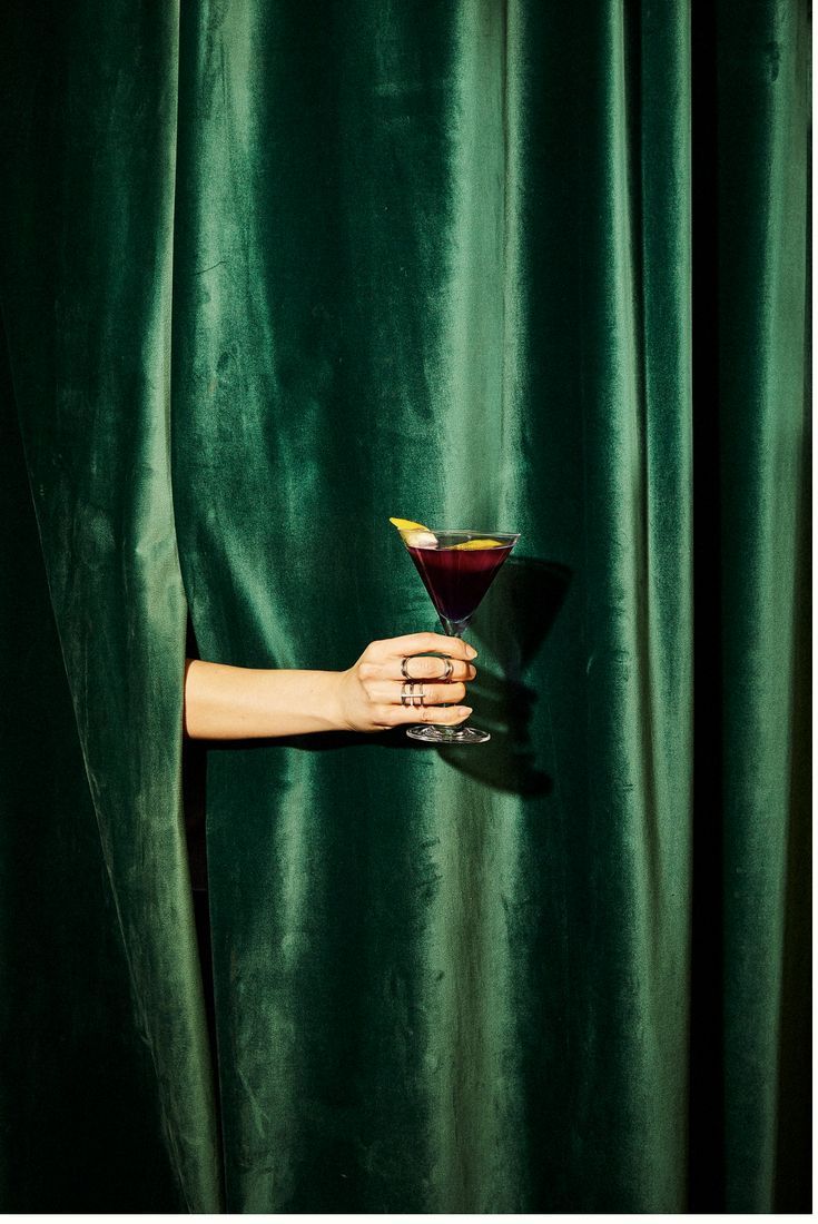 a woman holding a martini glass in front of a green curtain with her hand on it