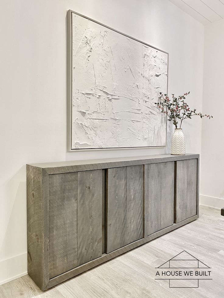 a white vase sitting on top of a wooden cabinet next to a wall mounted art piece