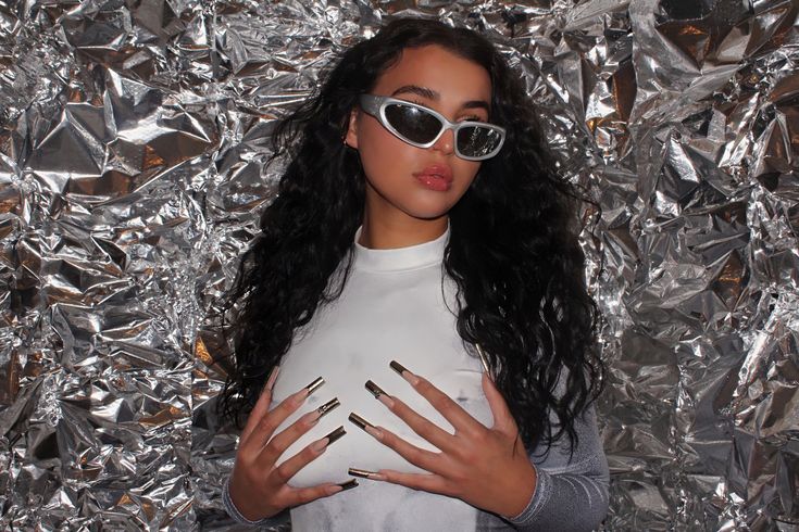 a woman with long black hair wearing sunglasses and holding her hands on her chest in front of silver foil