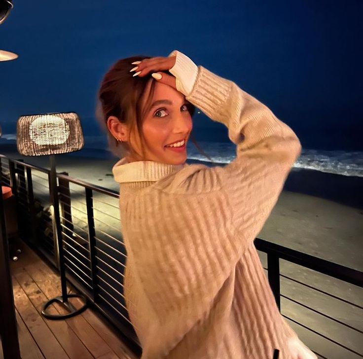 a woman standing next to a railing near the ocean at night with her hands on her head