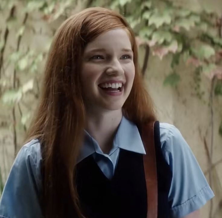 a girl with long red hair wearing a blue shirt and brown suspenders smiles at the camera