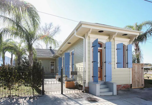 a small white house with blue shutters on the front and side doors, surrounded by palm trees