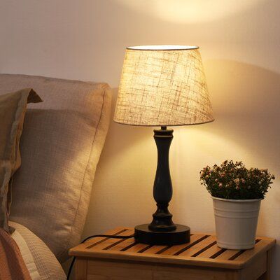 a lamp on a night stand next to a bed with pillows and a potted plant