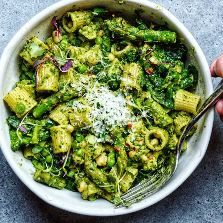 a white bowl filled with pasta and pesto covered in parmesan cheese next to a hand holding a fork