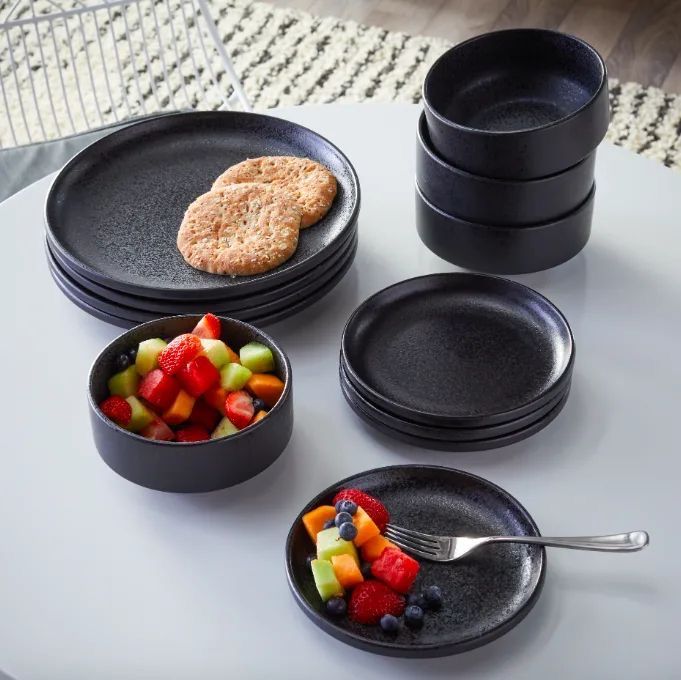 four black plates with fruit and crackers in them sitting on a white tablecloth