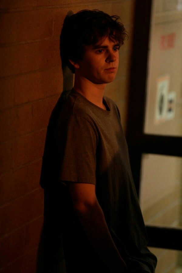 a young man standing in front of a window next to a brick wall at night