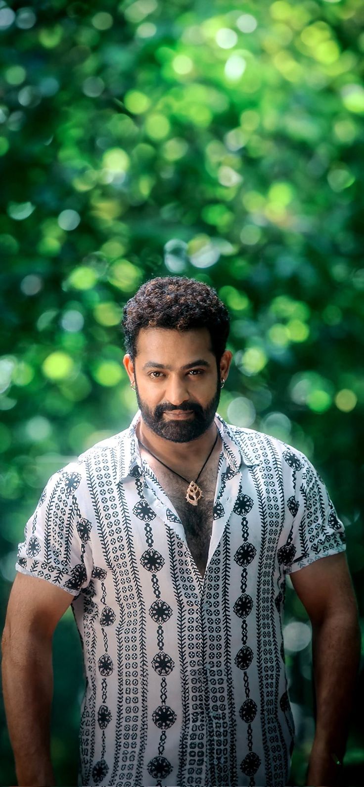 a man with a beard standing in front of green leaves and trees, looking at the camera