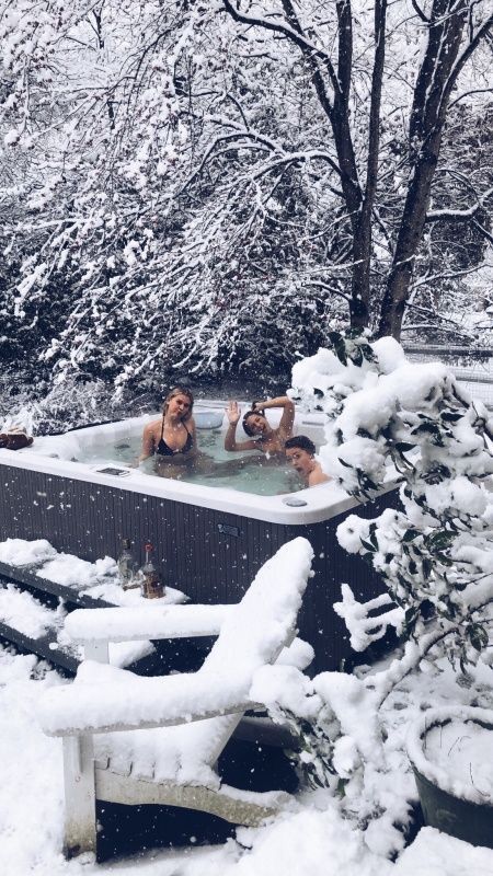 two people in a hot tub surrounded by snow covered trees and bushes, with one person taking a photo