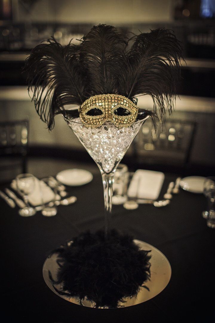 a masquerade mask sitting on top of a martini glass filled with black feathers