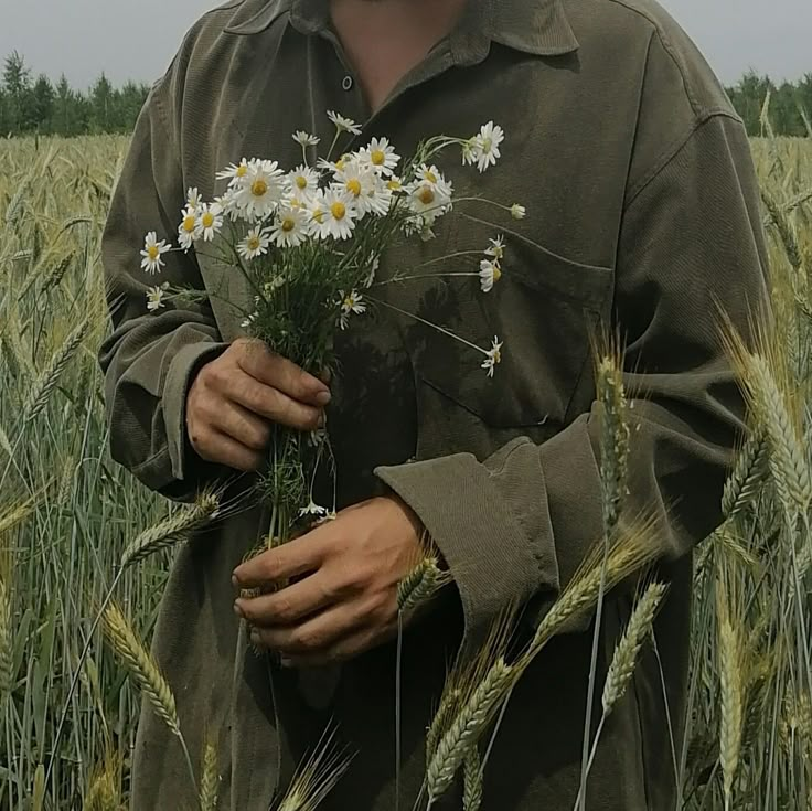 a man standing in a wheat field holding daisies and looking at the camera with an overcast sky behind him