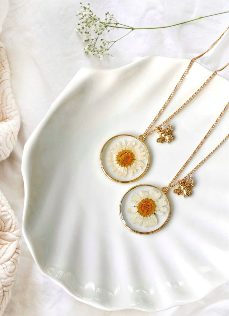 two necklaces with daisies on them sitting on a white plate next to dried flowers