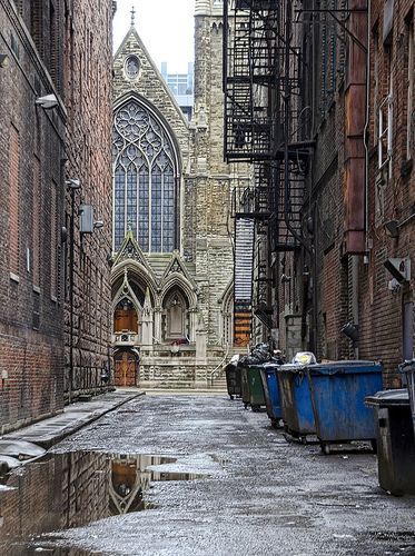 an alleyway with trash cans and buildings in the background, on a rainy day