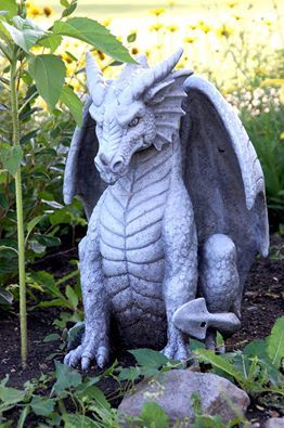 a statue of a dragon sitting on the ground in front of some plants and flowers