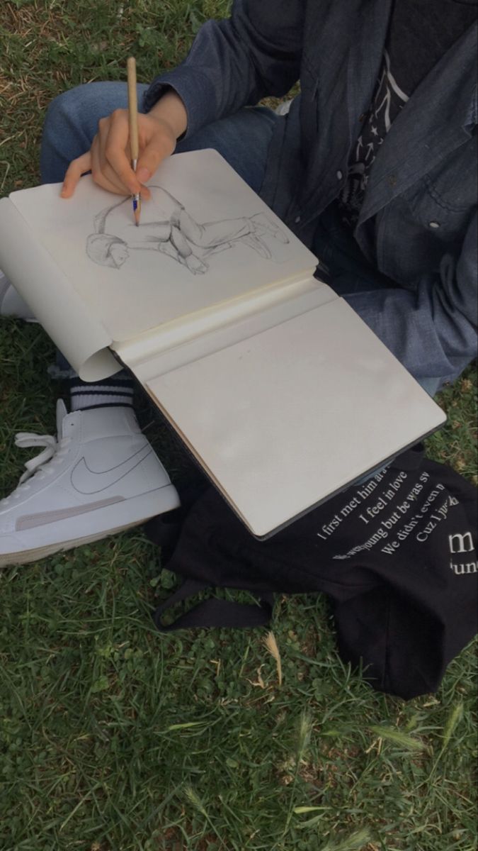 a person sitting in the grass drawing with a pencil and paper on top of it