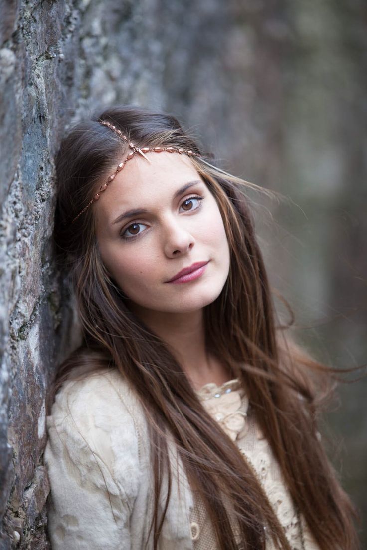 a woman with long hair standing next to a stone wall and looking at the camera