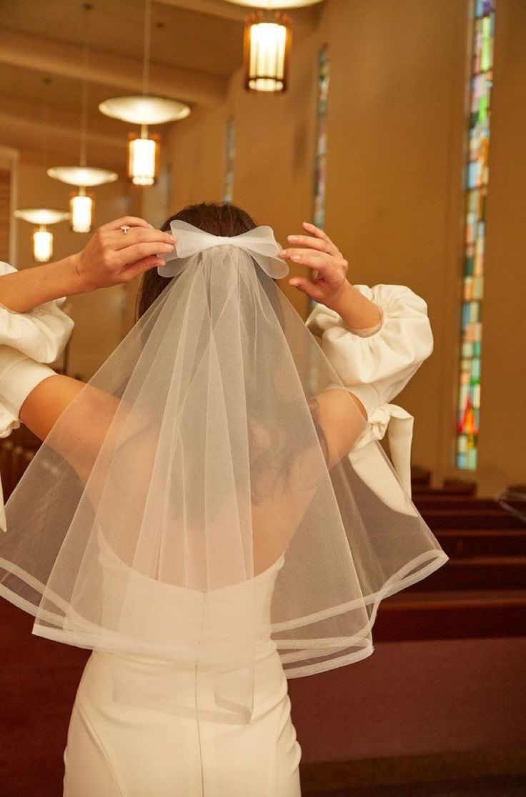 a woman in a white dress and veil covering her face with both hands while standing in front of a church pew