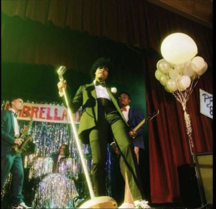 a man in a green suit on stage holding a microphone and standing next to balloons