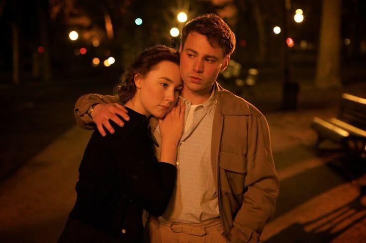 a man and woman standing next to each other on a sidewalk at night time with street lights in the background