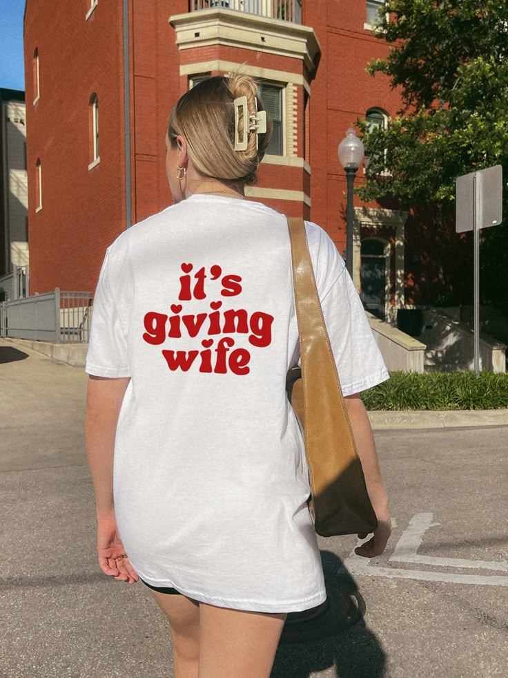 a woman walking down the street wearing a t - shirt that says it's giving wife