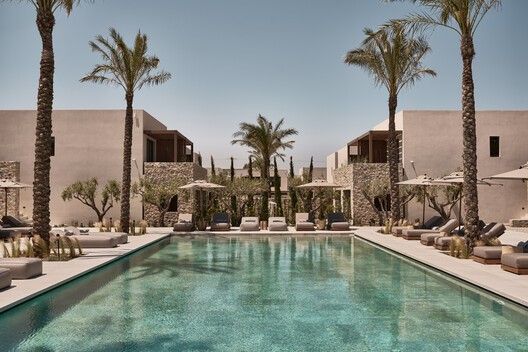 an outdoor swimming pool with lounge chairs and palm trees in the foreground, surrounded by two buildings