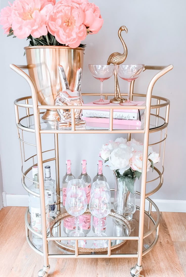 a gold metal bar cart with pink flowers and wine glasses on the bottom shelf next to it