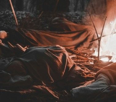 two people are sitting in front of a campfire while one person is laying on the ground